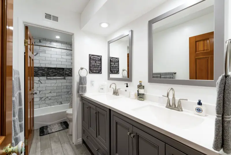 Modern bathroom remodel in Calabasas featuring a double gray vanity with white countertops, two gray-framed mirrors, and a tiled shower area seen through an open wood door.