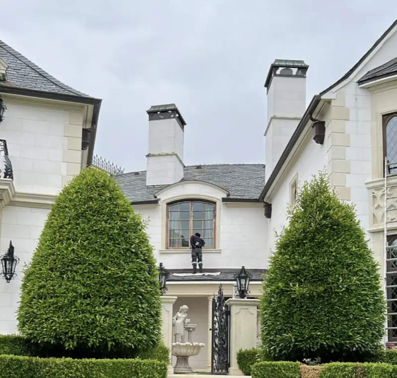 A professional worker cleaning the second-story exterior windows of a large, luxury white stone estate featuring manicured cone-shaped hedges and a green lawn.