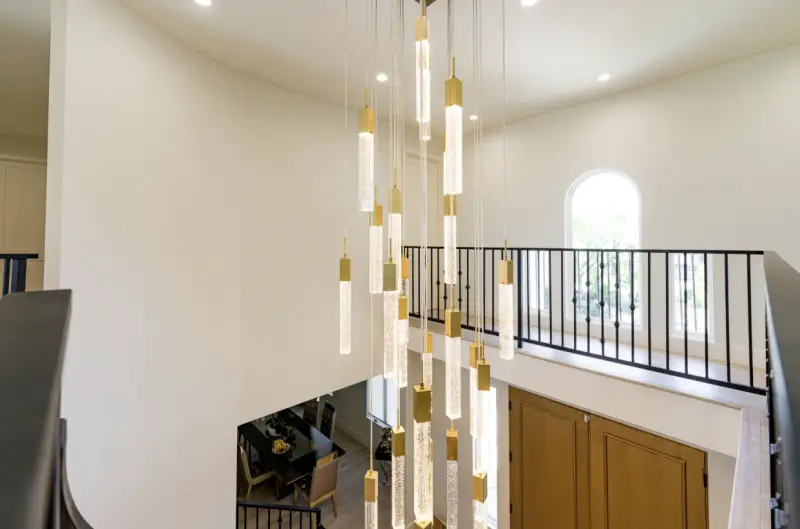 A modern, multi-light cascading chandelier with gold accents and cylindrical glass pendants hanging in a high-ceiling foyer above a dining area and staircase.