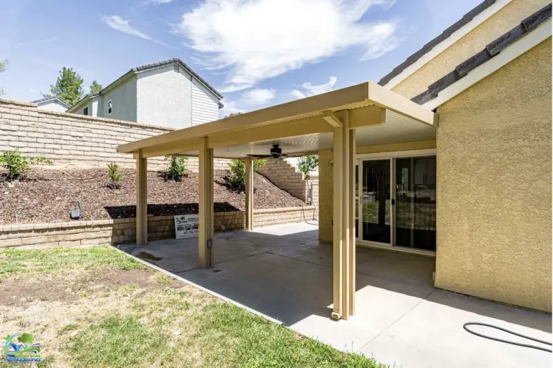 A solid tan aluminum patio cover with a ceiling fan installed over a concrete backyard patio, attached to a stucco home with sliding glass doors.