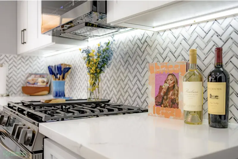 A modern kitchen remodel featuring a white herringbone tile backsplash, white marble countertops, and a stainless steel gas range with under-cabinet lighting.