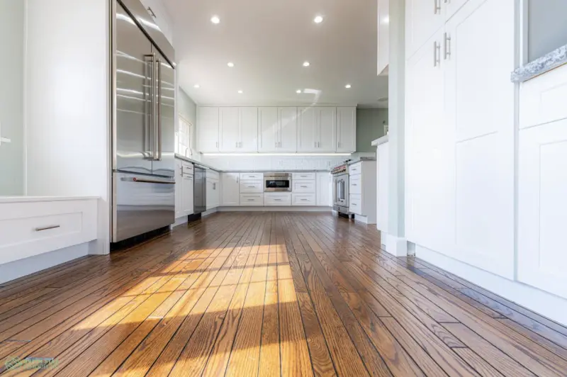 A bright, modern kitchen featuring white shaker cabinets, a large stainless steel refrigerator, recessed ceiling lighting, and polished dark hardwood flooring.