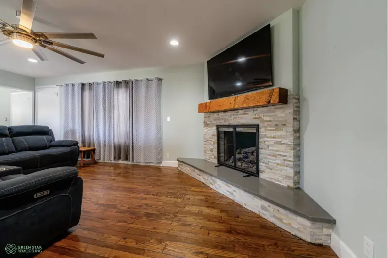 A modern living room featuring a stone-clad fireplace with a thick wood mantel, a wall-mounted television, and polished hardwood floors.