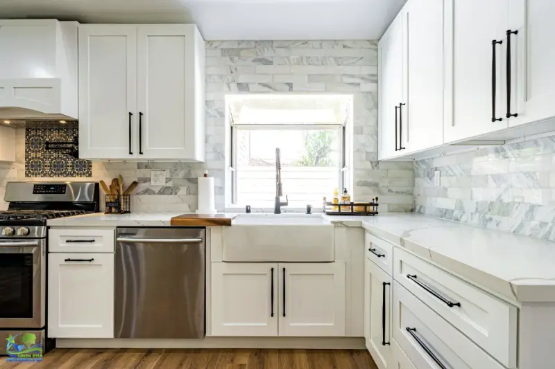 Modern kitchen featuring white shaker cabinets with black hardware, a large white farmhouse sink beneath a window, and light marble subway tile backsplash.