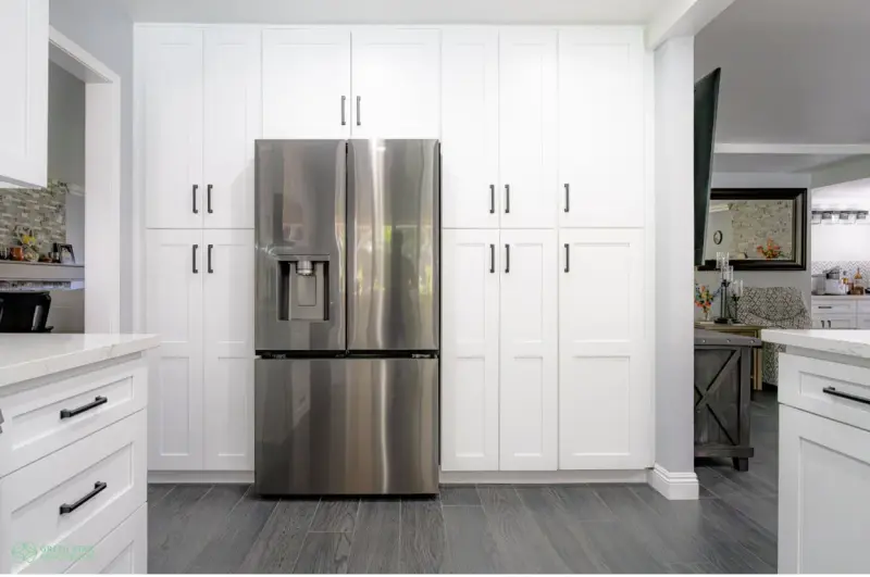 A modern kitchen featuring floor-to-ceiling white shaker-style pantry cabinets with matte black handles surrounding a built-in stainless steel French door refrigerator on dark gray wood-look tile flooring.