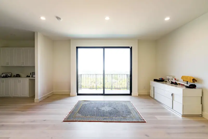 A bright, minimalist modern room featuring light wood flooring, a blue patterned area rug, a white dresser, and a large sliding glass door opening to a balcony with an outdoor view.