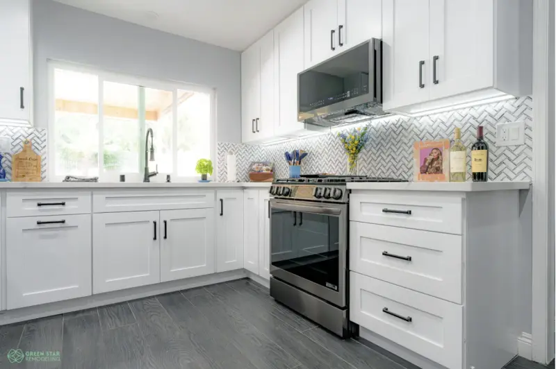 A modern kitchen remodel featuring white shaker cabinets with black hardware, a grey herringbone backsplash, stainless steel appliances, and dark wood-style flooring by Green Star.