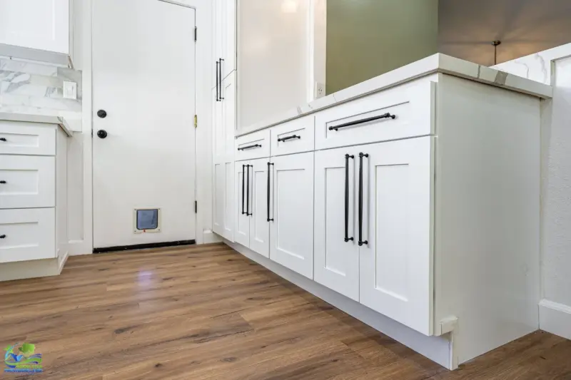 A modern kitchen featuring white shaker-style cabinets with matte black hardware, light brown hardwood flooring, and a white door with a pet flap.