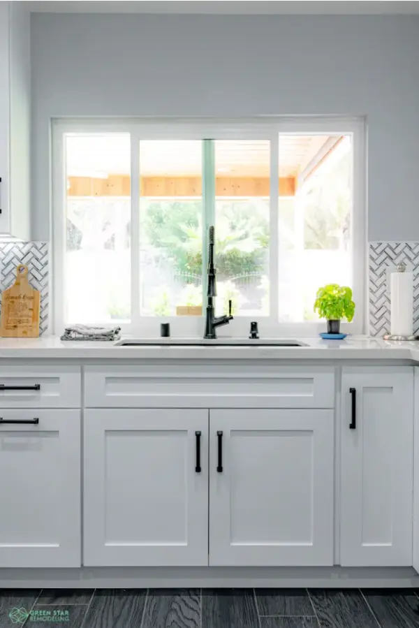 A modern kitchen remodel by Green Star Remodeling featuring white shaker-style cabinets with black hardware, a black farmhouse-style faucet, and a large window overlooking an outdoor patio.