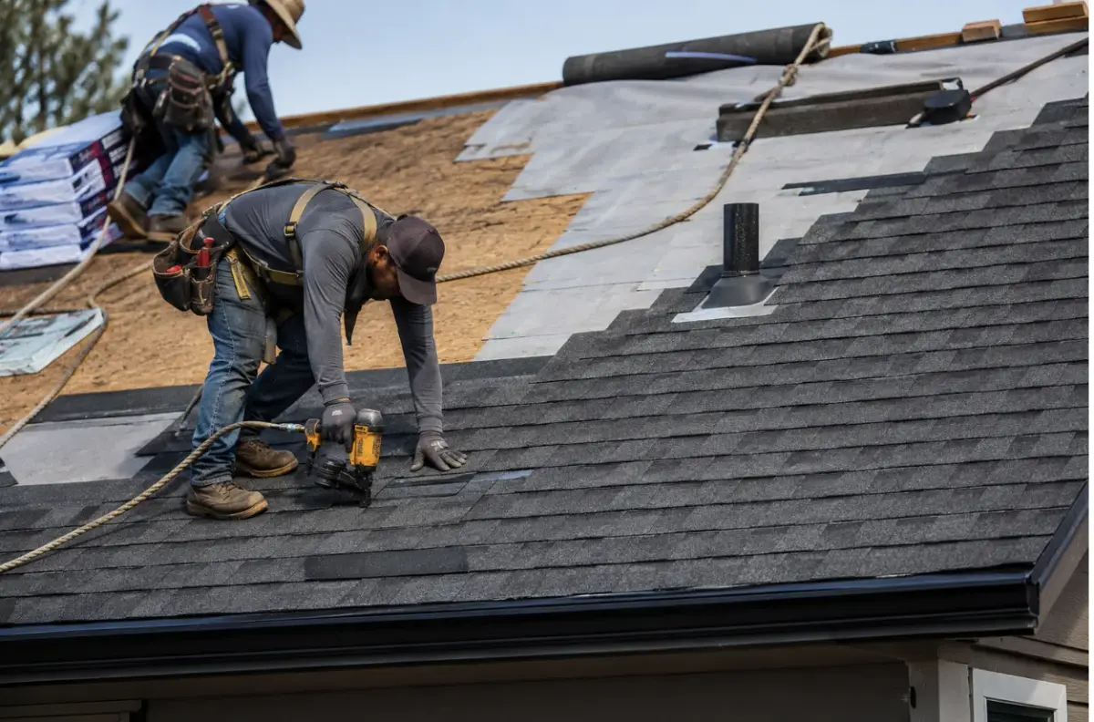 Professional roofing contractors installing architectural asphalt shingles over synthetic underlayment on a residential roof with roof vent flashing, drip edge, roofing nail gun, and completed shingle sections in bright daylight.
