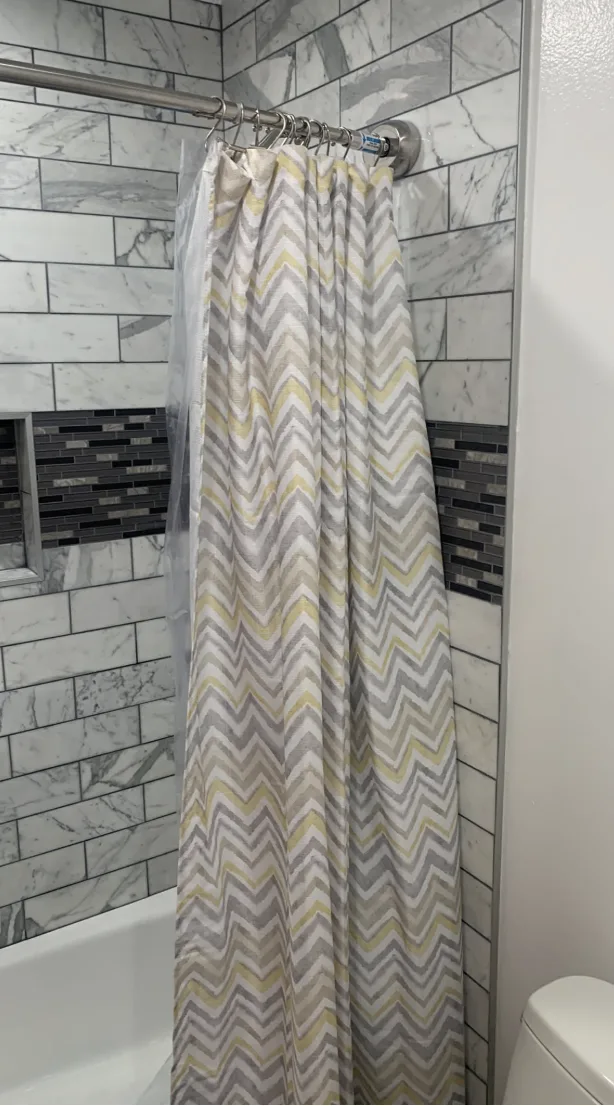 A modern shower curtain featuring a gray and yellow chevron zigzag pattern hanging in a bathroom with marble-style subway tiles and a dark accent tile border.