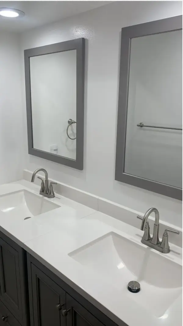 A modern bathroom featuring a double vanity with grey cabinets, white quartz countertops, integrated rectangular sinks, brushed nickel faucets, and two large grey-framed mirrors mounted on a white wall.