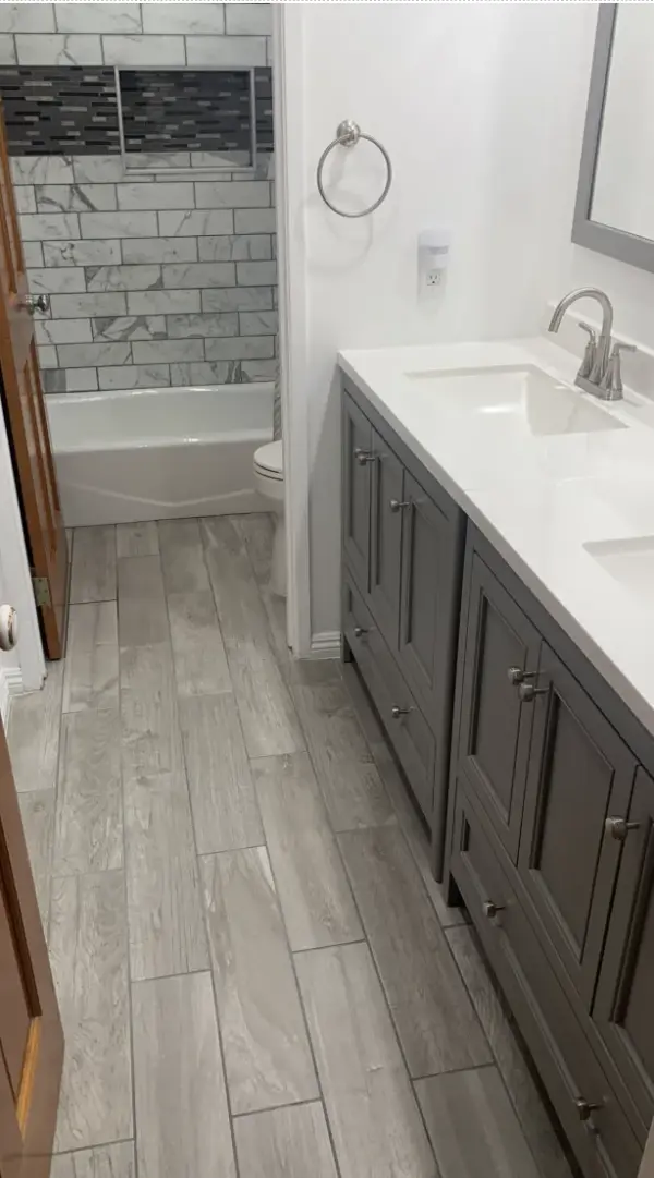 A modern bathroom featuring a double gray vanity with a white countertop, wood-look floor tiling, and a white marble-patterned subway tile shower with a dark mosaic accent niche.